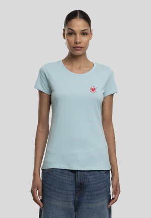 Femme aux cheveux foncés attachés, portant un t-shirt bleu clair avec un petit cœur rouge et un motif de rayons sur la poitrine gauche et un jean bleu.