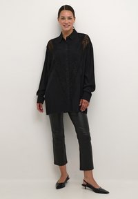 Camicia nera con bottoni e dettagli in pizzo sulle spalle, abbinata a pantaloni cropped in finta pelle neri e ballerine con punta.