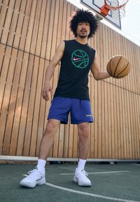 Fekete ujjatlan mez zöld és rózsaszín Nike logóval, kék hálós rövidnadrág arany "WARRIORS" felirattal, fehér sneaker, kosárlabdát tartva.