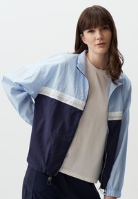 Giacca a vento blu chiaro e blu navy con striscia bianca, chiusura con zip, polsini elasticizzati e orlo regolabile. Indossata sopra una maglietta di colore neutro.