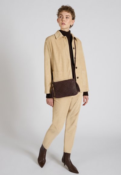 Chaqueta de pana beige, suéter de cuello alto marrón, pantalones de pana beige, bolso tejido marrón oscuro con borlas y botas de tobillo de punta afilada marrones.