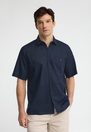 Man staat met handen gedeeltelijk in de zakken, draagt een marineblauw overhemd met korte mouwen en een klein rood logo op de borstzak.