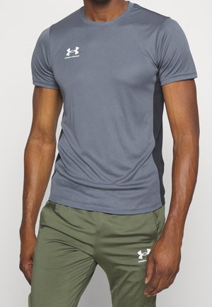 Torso masculin portant un t-shirt gris ajusté à manches courtes Under Armour et un pantalon vert olive Under Armour, debout devant un fond uni.