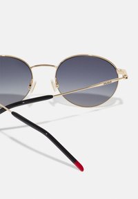 Ochelari de soare rotunzi cu lentile degradate care trec de la întunecat la deschis. Cadru din metal auriu cu brațe negre care au un vârf roșu, design minimalist.