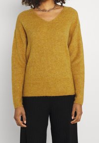 Musztardowy sweter w serek o prążkowanej fakturze, z obniżonymi ramionami i długimi rękawami. W połączeniu z czarnymi prążkowanymi spodniami.