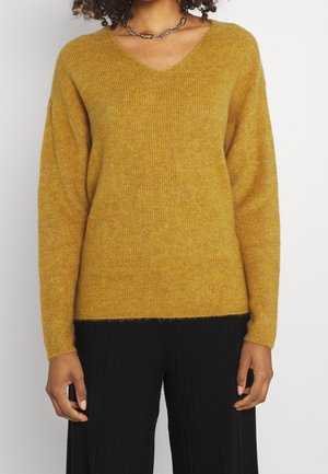 Mosterdgele v-hals trui met ribbelstructuur, verlaagde schouders en lange mouwen. Gecombineerd met zwarte ribbelbroeken.