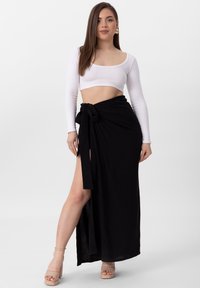 Czarna spódnica o kroju wrap z bocznym rozcięciem i zawiązywaniem w pasie, zestawiona z białą, długą bluzką typu cropped. Modelka nosi nagie, platformowe obcasy.
