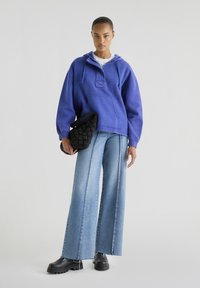 Overmaatse blauwe hoodie met een voorzak, gecombineerd met wijde denim jeans met een lichte wasbeurt en zichtbare stiksels, en een zwarte handtas met textuur.