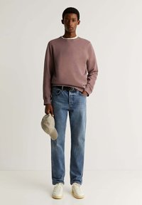 Felpa viola pallido, jeans dritti blu, sneakers bianche, che tiene in mano un cappellino grigio chiaro. Tessuto morbido e design con vestibilità rilassata.