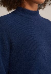 Marinblå, texturerad tröja med hög krage, tillverkad av mjukt, lurvigt material. Har en figursydd form och ribbat detaljer vid kragen.