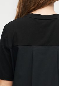 Άτομο με μακριά καστανά μαλλιά που φοράει μαύρο μπλουζάκι με κοντό μανίκι, με υφασμάτινη επένδυση στο άνω μέρος της πλάτης και λεπτομέρεια με πιέτα.