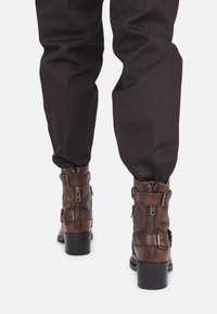 Botas de tobillo de cuero marrón con cremalleras, hebillas y punta redondeada, combinadas con pantalones oscuros, destacando el material texturizado y el diseño.
