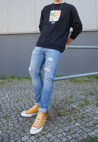 Persona apoyada contra una barandilla de metal, vistiendo un sudadera negra con gráficos, jeans light blue rasgados y zapatillas altas mostaza.