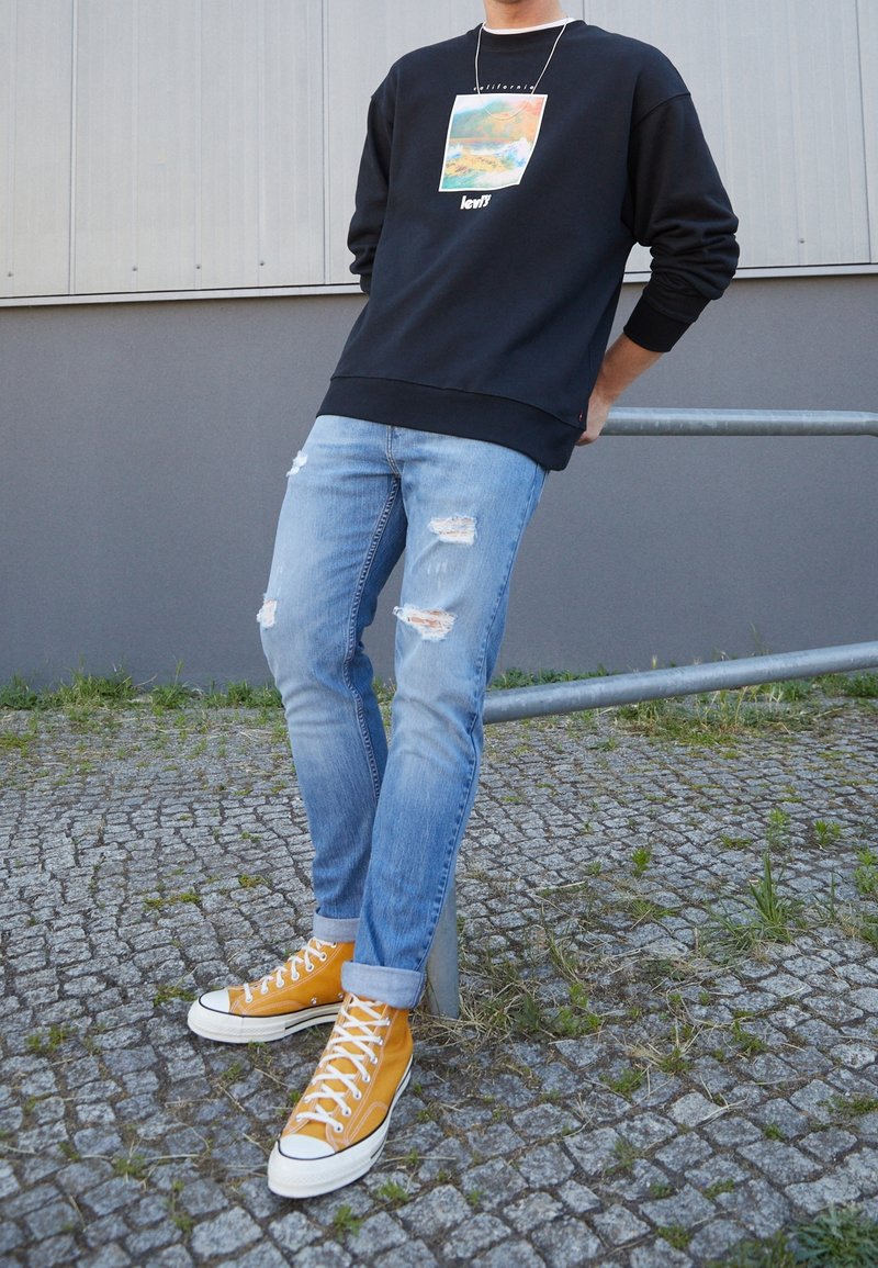 Persona apoyada contra una barandilla de metal, vistiendo un sudadera negra con gráficos, jeans light blue rasgados y zapatillas altas mostaza.