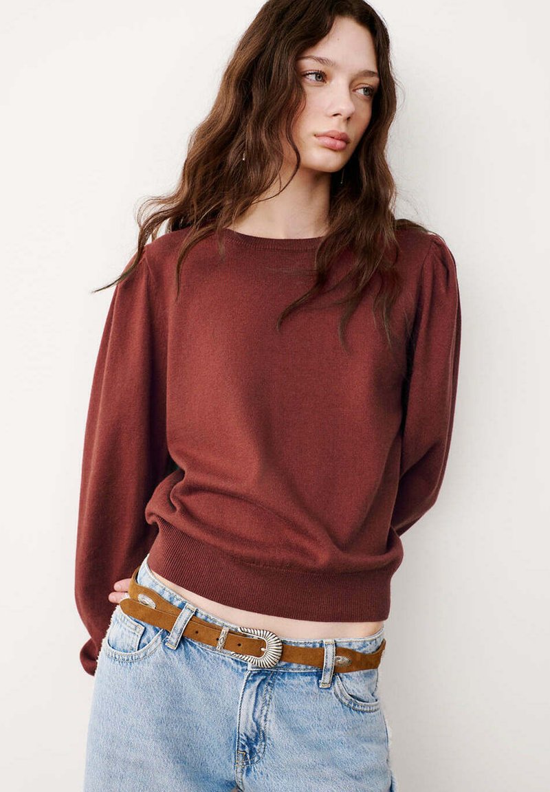 Jeune femme aux longs cheveux bruns portant un pull couleur rouille et un jean bleu clair avec une ceinture marron, regardant sur le côté sur un fond uni.