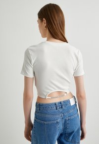 Top corto de manga corta blanco con relieve que presenta un pequeño corte en la parte de atrás con una banda decorativa. Combinado con unos jeans de mezclilla azul de talle alto.