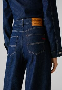 Jean en denim bleu foncé avec une patch en cuir portant l'inscription "Red Bull Racing x Pepe Jeans," avec deux poches arrière et des surpiqûres contrastées.