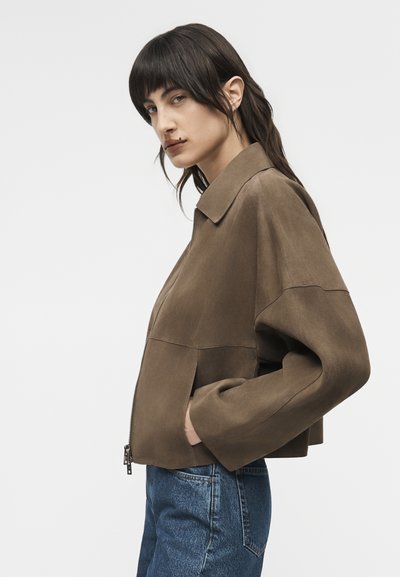 Γυναίκα με σκούρα μαλλιά φοράει καφέ καφέ μπουφάν από σουέτ και μπλε τζιν, με τα χέρια στις τσέπες του μπουφάν, αντικρίζοντας προς τα αριστερά σε μια απλή φόντο.