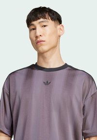 Fialové sportovní tričko s vertikálními pruhy, kulatým výstřihem a černými detaily. Obsahuje malé černé logo v oblasti hrudníku. Hladká, lehká tkanina.