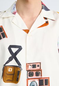 Camisa de manga corta y botones de tela crema con estampados gráficos de varios objetos, incluyendo una bolsa, altavoces y ventanas en colores vibrantes.