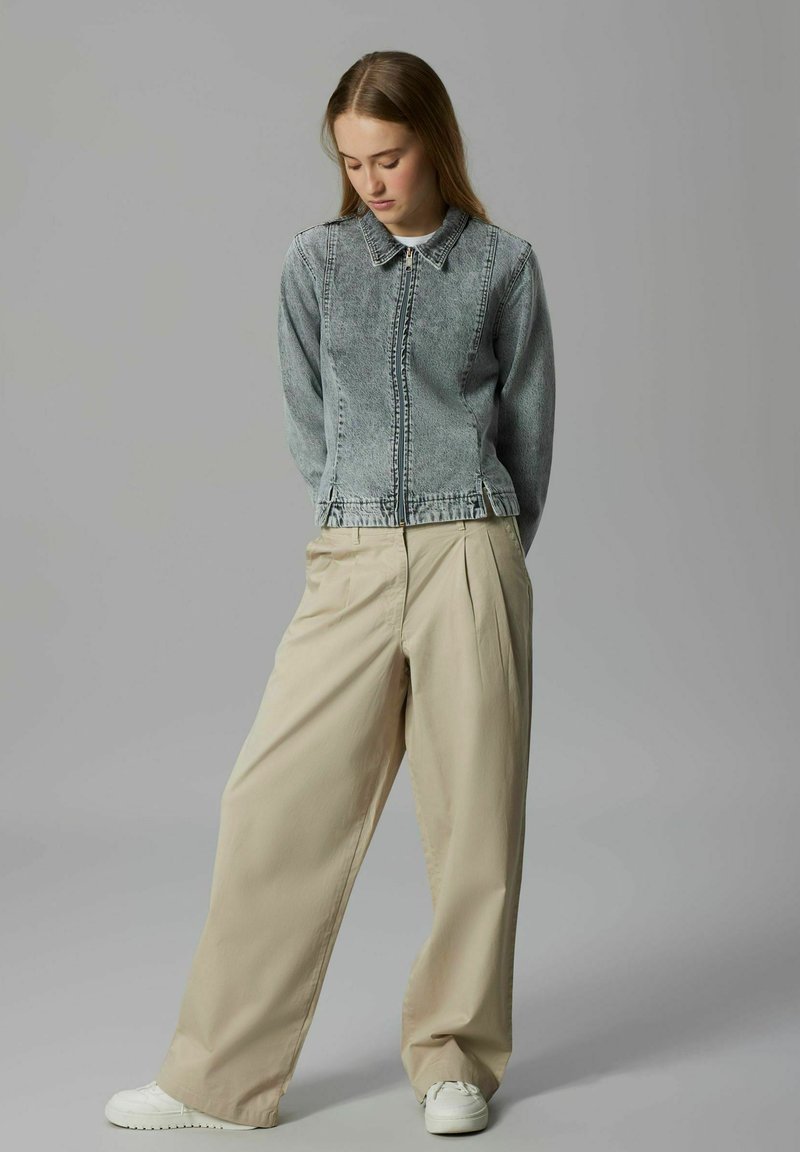 Jeune femme regardant vers le bas, portant une veste en denim bleu délavé, un pantalon large beige et des baskets blanches, posant devant un fond gris.