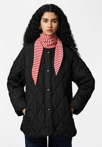 Manteau noir matelassé avec une coupe carrée, doté de boutons-pression. Associé à une écharpe rayée rouge et blanche, créant un contraste de couleurs.