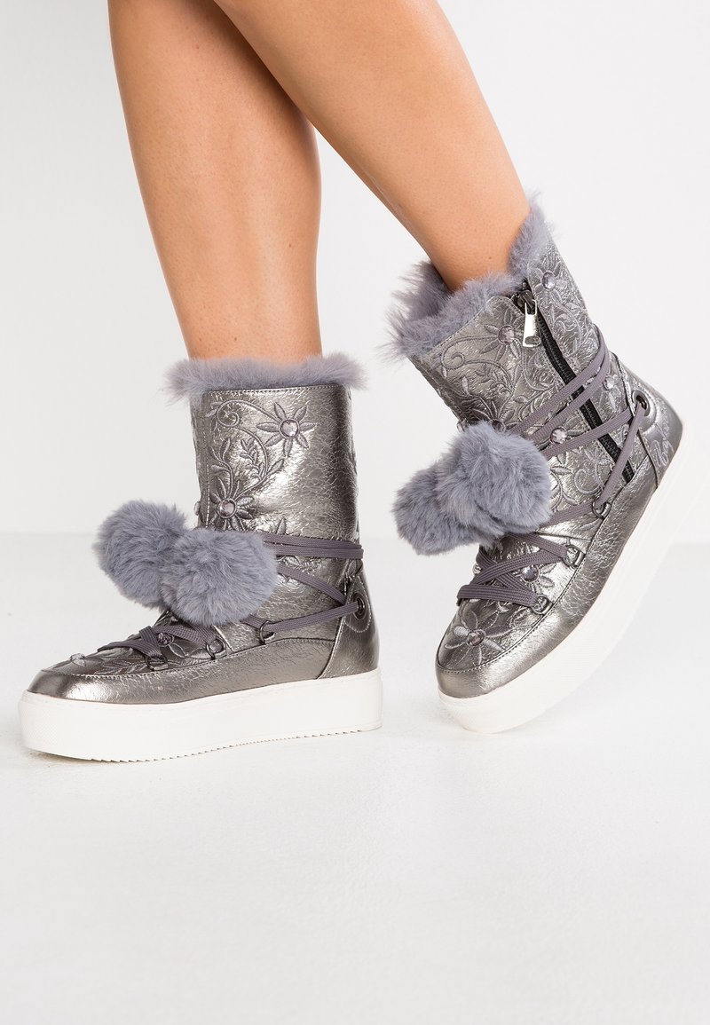 Bottes mi-mollet argentées métalliques avec broderie florale, garniture en fausse fourrure grise et pompons, fermeture éclair sur le côté et semelles en plateforme blanches portées sur des jambes.