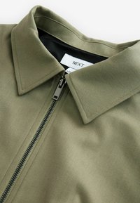 Olivgrüne Jacke mit Fischgrätstruktur, ausgestattet mit einem schwarzen Reißverschluss und einem spitzen Kragen. Das Etikett innen am Kragen trägt die Aufschrift "NEXT".