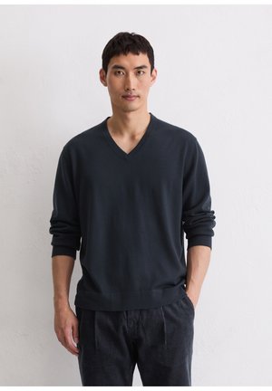 Sweater de gola V azul-marinho com mangas longas, tecido macio, punhos e bainha canelados. Usado com calças de veludo cotelê escuras. Fundo neutro.
