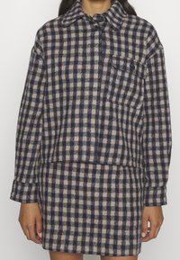 Chemise en flanelle à carreaux bleu marine et beige avec un col, fermeture à boutons et une poche poitrine, associée à une jupe assortie. Tissu texturé.