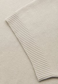 Tejido de suéter de color beige claro que muestra una superficie texturizada con un detalle de puño acanalado. El diseño general es simple y minimalista.