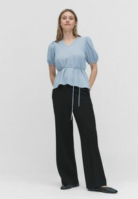 Ljusblå blus med puffärmar, V-ringning och justerbart midjeband, kombinerad med högmidjade svarta byxor och svarta platta skor.