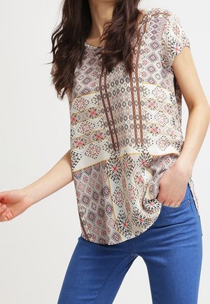 Kvinde iført en kortærmet bluse med geometrisk mønster i lyserøde, sorte og beige nuancer, kombineret med blå jeans, hånd i lommen.
