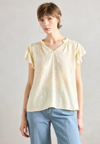 Lys gul bluse med korte volangermmer, V-hals og subtilt teksturert m�ønster. Matcher med lyseblå, løstsittende denimjeans.