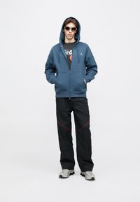 Sweat à capuche bleu zippé avec capuche, pantalon cargo noir avec accents rouges, et baskets grises. Le mannequin porte des lunettes de soleil et un t-shirt graphique.