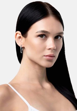 Orecchino a cerchio in argento con finitura lucida, indossato da una persona con capelli lunghi e scuri e tono della pelle neutro, su sfondo grigio chiaro.