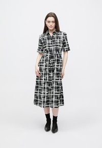 Vestido camiseiro aos quadrados preto e branco, com mangas curtas, cintura franzida e comprimento até ao meio da perna, combinado com botas tipo meias pretas.