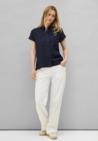 Camicia blu navy a maniche corte con chiusura a bottoni e due tasche sul petto, abbinata a pantaloni larghi di colore bianco sporco e scarpe beige.