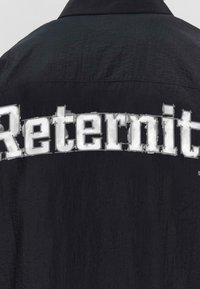 Schwarze Jacke aus strukturiertem Stoff mit großen, weißen gedruckten Buchstaben "Reternit" auf dem Rücken.