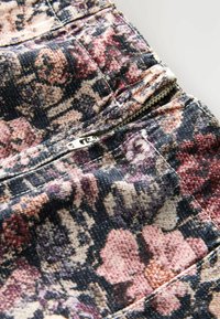 Ziedu raksts audumā rozā, purpura un melnā krāsā, tuvplānā redzama rāvējpulcējs detalizācija biksēs. Strukturēts džinsu materiāls.