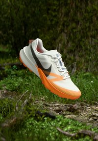 Fehér hálós Nike futócipő narancssárga és fekete részletekkel, texturált felülettel, könnyű kialakítással és látható fűzővel.