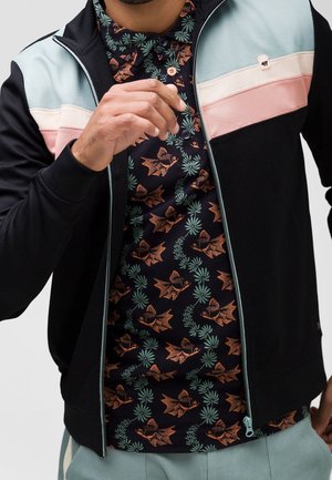 Man draagt een zwarte jas met rits met lichtblauwe en roze strepen over een zwart shirt met patroon van oranje vissen en groene zeewier.