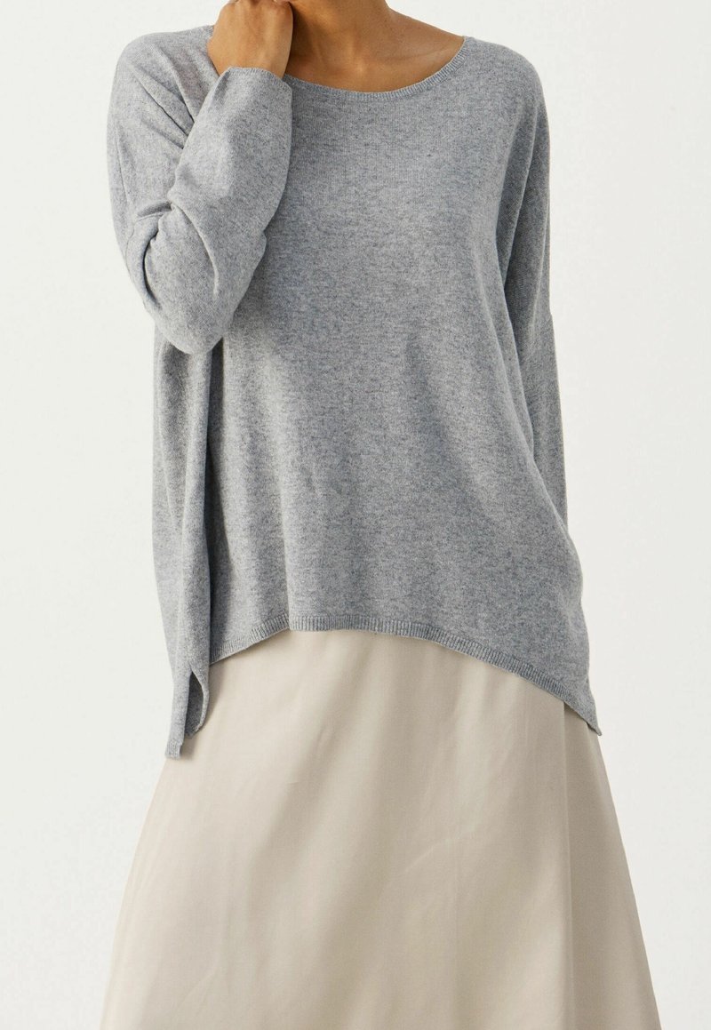 Femme portant un pull gris ample à manches longues avec des fentes latérales et une jupe beige sur un fond clair uni.