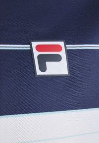 Námořnická textilie s horizontálními pruhy, obsahující významné logo FILA v červené a šedé barvě, umístěné na bílém a světle modrém pozadí.