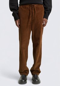 Braune Cordhose mit lockerem Schnitt, ausgestattet mit einem Tunnelzugbund und sichtbarer vertikaler Rippenstruktur. Kombiniert mit schwarzen Stiefeln.