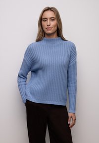 Pull tricot bleu clair avec un col montant, une texture côtelée et des manches longues. Associé à un pantalon marron foncé, avec une coupe décontractée.