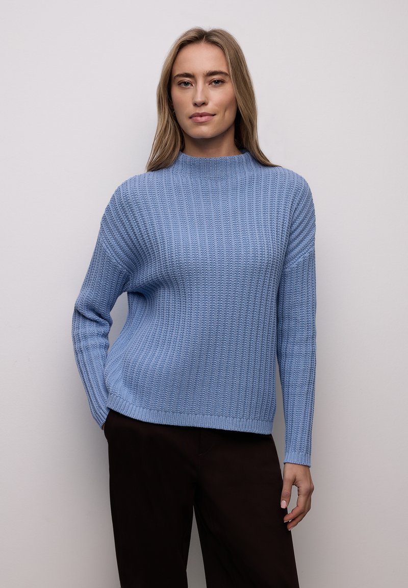 Pull tricot bleu clair avec un col montant, une texture côtelée et des manches longues. Associé à un pantalon marron foncé, avec une coupe décontractée.