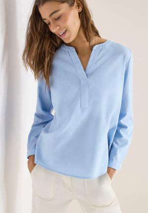 Joven sonriente, vestida con una blusa de manga larga color azul claro con textura y pantalones color blanco roto, con las manos en los bolsillos, sobre un fondo liso.