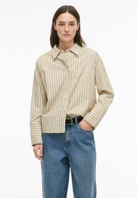 Chemise beige à rayures avec boutons sur le devant, col et manches longues. Associée à un jean denim bleu taille haute et une ceinture noire.