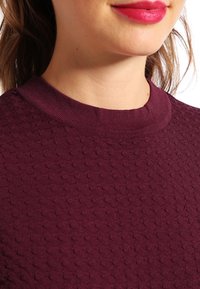 Pull en maille bordeaux avec un motif texturé, doté d'un col rond et de manches courtes. La vue rapprochée met en avant les détails de son tissu.
